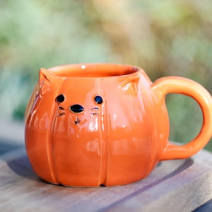 May include: An orange ceramic mug in the shape of a pumpkin with cat features. The mug has a round body with vertical ridges, a curved handle, and small ears. Black dots and lines form the cat's eyes, nose, and whiskers. The mug is on a wooden surface.