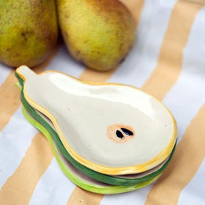 May include: A stack of pear-shaped ceramic dishes in shades of white, yellow, and green, with a pear design. Two ripe pears are in the background on a striped cloth. The dishes are stacked on top of each other.