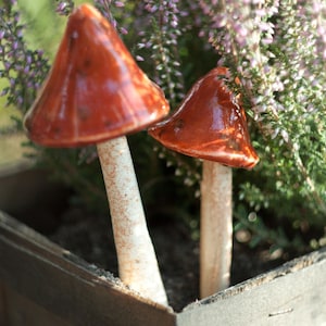 Peut inclure: Deux sculptures de champignons en céramique avec des chapeaux rouge-orange brillants et des tiges blanc cassé texturées. Les champignons sont placés à la verticale, le plus grand à gauche et un plus petit à droite, avec des surfaces lisses.
