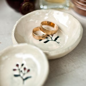 Może przedstawiać: Dwie złote obrączki spoczywają w małej, kremowej ceramicznej miseczce z namalowanym botanicznym wzorem. Kolejna podobna miseczka znajduje się na pierwszym planie. Tło obejmuje ciemnobrązową butelkę i przezroczystą butelkę.