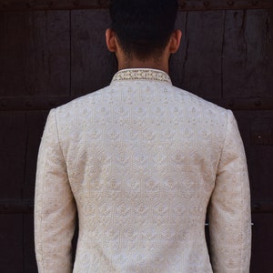 Puede incluir: Chaqueta bordada color crema con cuello mandar&iacute;n. La chaqueta presenta intrincados patrones florales y geom&eacute;tricos. Las mangas son largas y la chaqueta parece estar hecha de una tela texturizada. El fondo es una puerta de madera marr&oacute;n oscuro.