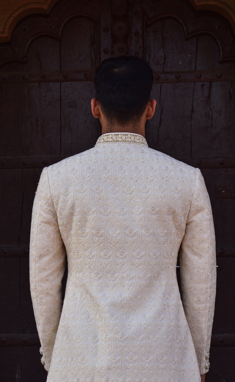 Puede incluir: Chaqueta bordada color crema con cuello mandar&iacute;n. La chaqueta presenta intrincados patrones florales y geom&eacute;tricos. Las mangas son largas y la chaqueta parece estar hecha de una tela texturizada. El fondo es una puerta de madera marr&oacute;n oscuro.