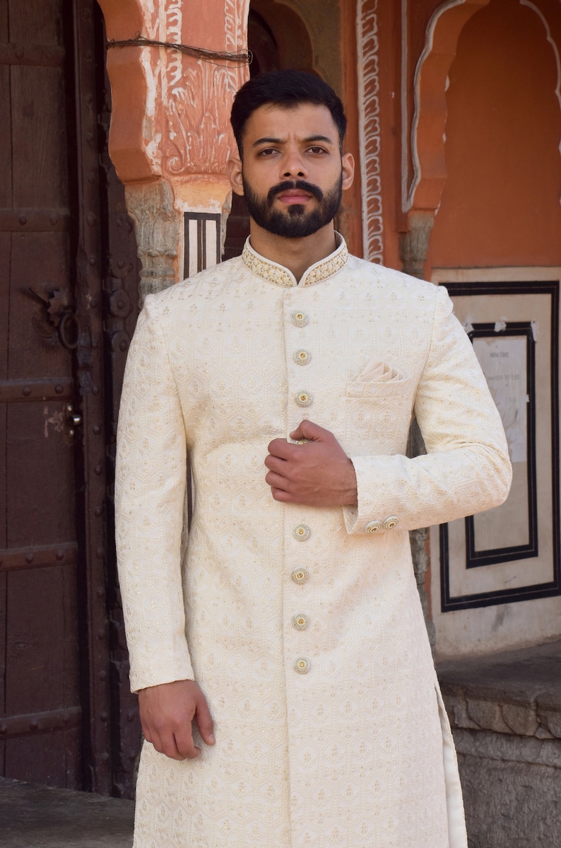 Puede incluir: Sherwani bordado de color marfil con cuello mandar&iacute;n y botones dorados. El abrigo largo y a medida presenta un pa&ntilde;uelo de bolsillo y detalles intrincados, adecuado para ocasiones formales. La prenda es ropa formal tradicional del sur de Asia.