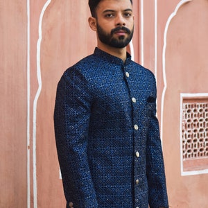 K&ouml;nnte beinhalten: Marineblaue bestickte Jacke mit geometrischem Muster und goldenen Kn&ouml;pfen. Die Jacke hat einen Stehkragen und ein Einstecktuch. Der Hintergrund ist eine rosa Wand mit architektonischen Details.