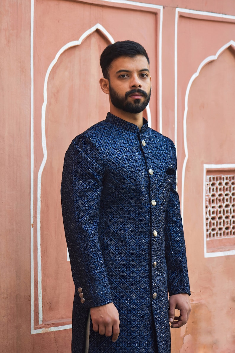 K&ouml;nnte beinhalten: Marineblaue bestickte Jacke mit geometrischem Muster und goldenen Kn&ouml;pfen. Die Jacke hat einen Stehkragen und ein Einstecktuch. Der Hintergrund ist eine rosa Wand mit architektonischen Details.