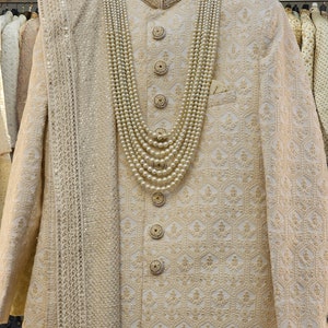 Puede incluir: Traje sherwani bordado de color crema con una bufanda a juego y un collar de perlas. La chaqueta presenta bordados dorados ornamentados, botones decorativos y un cuello mandar&iacute;n. El collar de perlas es en capas.