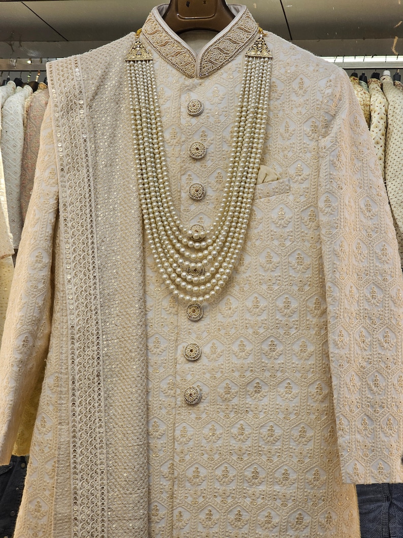 Puede incluir: Traje sherwani bordado de color crema con una bufanda a juego y un collar de perlas. La chaqueta presenta bordados dorados ornamentados, botones decorativos y un cuello mandar&iacute;n. El collar de perlas es en capas.