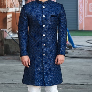 K&ouml;nnte beinhalten: Ein Mann tr&auml;gt eine lange, marineblaue Jacke mit Rautenmuster und wei&szlig;en Hosen. Die Jacke hat einen Stehkragen und silberne Kn&ouml;pfe. Das Outfit wird durch wei&szlig;e Schuhe erg&auml;nzt.