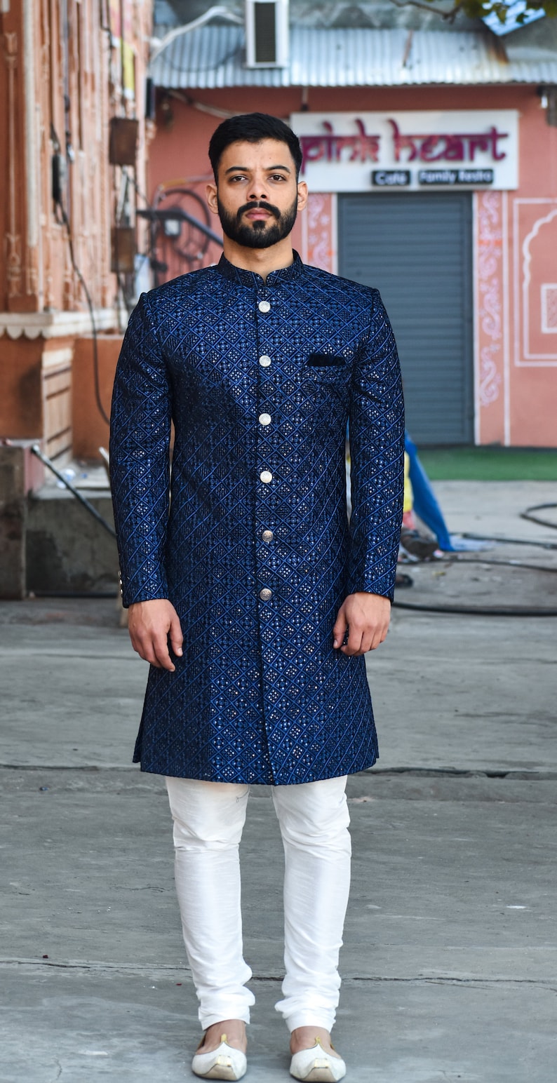 K&ouml;nnte beinhalten: Ein Mann tr&auml;gt eine lange, marineblaue Jacke mit Rautenmuster und wei&szlig;en Hosen. Die Jacke hat einen Stehkragen und silberne Kn&ouml;pfe. Das Outfit wird durch wei&szlig;e Schuhe erg&auml;nzt.