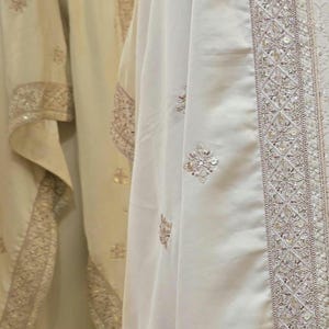 Könnte beinhalten: Ein weißer Schal mit aufwendigen Stickereien und Perlenbesatz. Der Schal ist aus einem weichen, fließenden Stoff gefertigt und hat eine zarte, feminine Ausstrahlung.