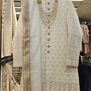 May include: Ivory-colored embroidered sherwani with gold accents, featuring intricate patterns and a matching scarf. The garment is adorned with decorative buttons and a pearl necklace. The sherwani is displayed on a hanger, showcasing its formal design.