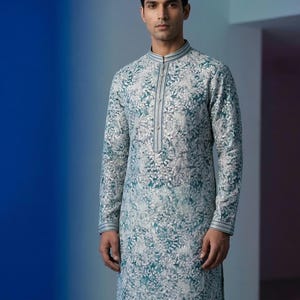 May include: A teal and white floral-patterned tunic with long sleeves, worn with white trousers. The tunic has a mandarin collar and a central stripe detail. The outfit is worn by a person. The outfit is suitable for formal occasions.