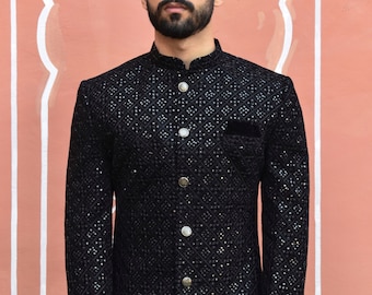 Könnte beinhalten: Schwarze Jacke mit geometrischem Muster und silbernen Knöpfen. Die Jacke hat einen Stehkragen und eine kleine Tasche. Der Hintergrund ist eine pfirsichfarbene Wand mit einem weißen dekorativen Bogen.