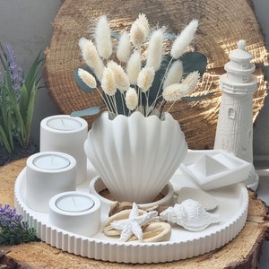 Könnte beinhalten: Weiße Keramikschale mit Muschel-Design, mit drei weißen Kerzenhaltern, einer weißen Muschelvase mit getrocknetem weißen Pampasgras, einer weißen Muschel, einem weißen Seestern und einer weißen Leuchtturmfigur.