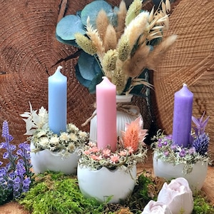 Può includere: Tre portacandele in ceramica bianca con candele blu, rosa e viola. Ogni portacandele è decorato con fiori secchi e verde.