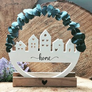 Op de afbeelding: Witte houten home decor met een uitsnijding van een stadsbeeld en het woord "home" in cursief. De decoratie is omringd door een krans van eucalyptusbladeren en staat op een houten voet.