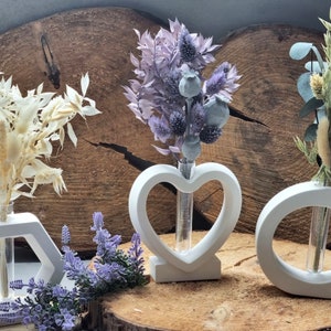 Puede incluir: Cuatro jarrones de cerámica blanca con tubos de vidrio para contener flores secas. Los jarrones tienen forma de hexágono, corazón y dos círculos. Las flores secas son blancas, moradas y verdes.