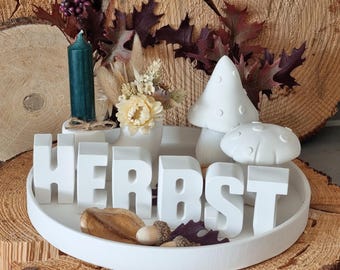 May include: White ceramic tray with the word "HERBST" spelled out in large white letters. The tray also features a small white ceramic mushroom, a green candle, and dried flowers.
