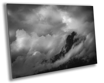 May include: A black and white photograph of a mountain peak partially obscured by clouds. The clouds are thick and billowing, creating a dramatic and moody atmosphere.