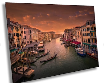 May include: A panoramic view of a canal in Venice, Italy, at sunset. The canal is lined with buildings on both sides, and there are several gondolas and boats in the water. The sky is a vibrant orange and pink, and the water is a deep blue.