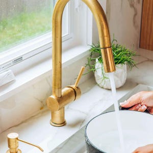 Può includere: Un rubinetto da cucina dorato con un becco ad arco alto è aperto, riempiendo una pentola nera d'acqua. Un dispenser di sapone dorato si trova sul bancone a sinistra del rubinetto.