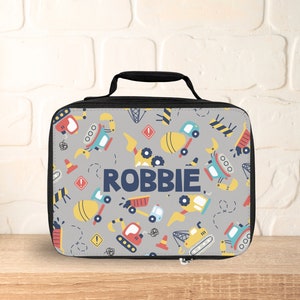 May include: A gray lunch bag with a black handle and zipper. The bag has a pattern of construction vehicles and the name "ROBBIE" printed in blue.