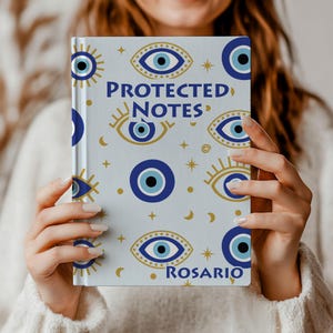 Könnte beinhalten: Ein hellblaues Notizbuch mit einem sich wiederholenden Muster aus blauen und goldenen Evil-Eye-Designs. Das Cover zeigt den Text "PROTECTED NOTES" und den Namen "ROSARIO". Das Notizbuch wird von einer Person gehalten, die einen weißen Pullover trägt.