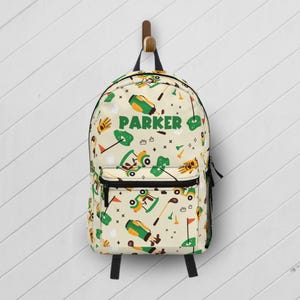 May include: A cream-colored backpack featuring a repeating pattern of golf-themed illustrations including golf carts, clubs, bags, and balls. The name PARKER is printed in bold green letters across the center of the front pocket area.