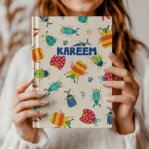 May include: A hardcover notebook with a cream-colored cover featuring a repeating pattern of colorful beetles and small floral icons. The name KAREEM is printed in bold blue capital letters across the center of the front cover.