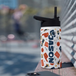 Könnte beinhalten: Eine weiße Wasserflasche mit schwarzem Deckel und Strohhalm. Die Flasche hat ein Basketball-Motiv mit dem Namen "JASON" in Blau und Weiß.