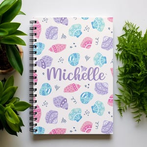 May include: A white spiral notebook with a colorful seashell pattern and the name "Michelle" written in purple.