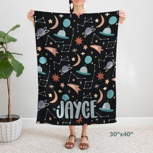 May include: A black fleece blanket with a space-themed pattern featuring planets, stars, and constellations. The name "JAYCE" is printed in white on the blanket. The blanket measures 30 inches by 40 inches.