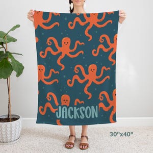 Könnte beinhalten: Ein blaugrünes Strandtuch mit einem sich wiederholenden Muster aus orangefarbenen Oktopus-Illustrationen. Das Handtuch trägt den Namen "JACKSON" in hellblauer Schrift. Das Handtuch misst 76 cm x 102 cm.