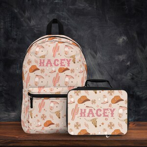 Op de afbeelding: Een roze en oranje gepatroneerde rugzak en lunchbox set met de naam "Kacey" op beide. Het patroon bevat cowboylaarzen, hoeden, cactussen en sterren.