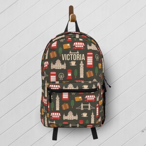 May include: A green backpack with a London theme. The backpack has a red, white, and blue pattern with images of double-decker buses, phone booths, Big Ben, and the London Eye. The name "VICTORIA" is printed in white on the front of the backpack.