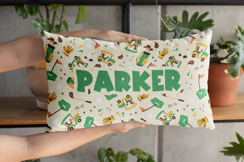 K&ouml;nnte beinhalten: Rechteckiges Kissen mit einem Golf-Motiv. Das Design umfasst Golfwagen, Golfschl&auml;ger, Golfb&auml;lle und den Namen "PARKER" in gro&szlig;en gr&uuml;nen Buchstaben. Der Hintergrund ist hellbeige.
