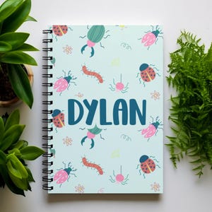 May include: A light blue spiral notebook with a colorful bug pattern. The notebook has the name "DYLAN" printed in dark blue letters.