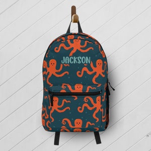 May include: A blue backpack with a repeating pattern of orange octopus illustrations. The backpack has a black zipper and a name tag that says "JACKSON".