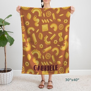 May include: A brown throw blanket with a repeating pattern of various pasta shapes in yellow and orange. The blanket has the name "Gabriele" printed in purple on the front. The blanket measures 30 inches by 40 inches.