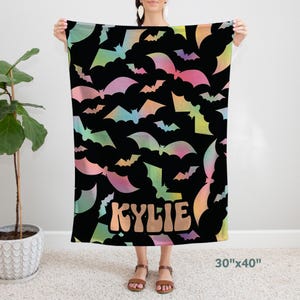 May include: A black fleece blanket with a colorful bat pattern and the name "KYLIE" in a retro font. The blanket measures 30 inches by 40 inches.