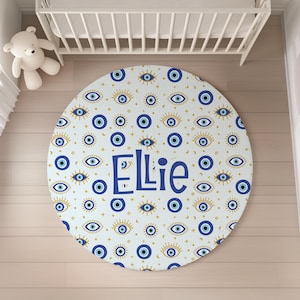 May include: A round, white play mat with a blue and gold pattern of evil eyes and stars. The name "Ellie" is written in blue in the center of the mat.