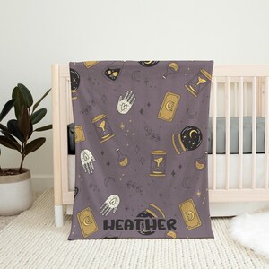 May include: Purple fleece blanket with a repeating pattern of gold and black illustrations of hands, moons, stars, hourglasses, and potion bottles. The blanket has the name "HEATHER" printed in black letters on the bottom.