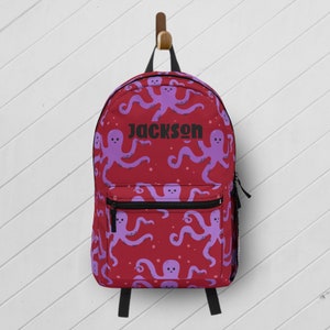 May include: A red backpack featuring a repeating pattern of light purple octopuses with small black eyes and tentacles. The name JACKSON is printed in bold black capital letters across the center of the bag. It includes a front zippered pocket.