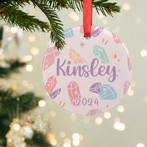May include: A pink and white Christmas ornament with a colorful crystal pattern and the name "Kinsley" and the year "2024" printed on it.
