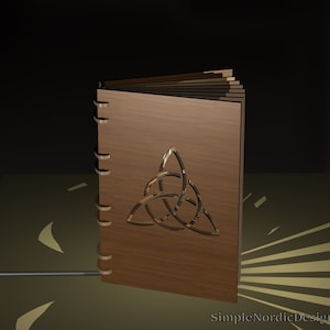 Puede incluir: Un libro de madera con un diseño de nudo celta tallado en la portada. El libro está cerrado y tiene un lomo con anillos metálicos.