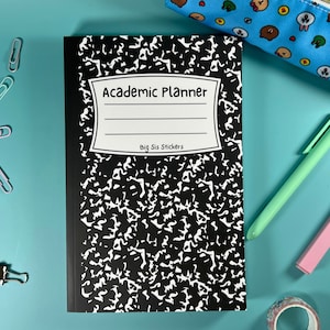 Peut inclure: Un cahier à couverture noire et blanche avec le texte "Academic Planner" et "Big Sis Stickers" sur la couverture. Le cahier est sur une surface bleue avec un crayon, un stylo et d'autres fournitures scolaires.