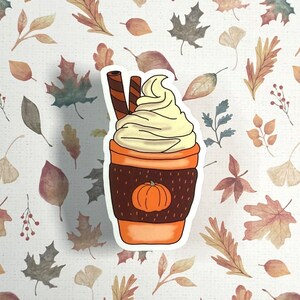 May include: A sticker of a pumpkin spice latte with whipped cream and a cinnamon stick. The latte is in a brown cup with a pumpkin design.