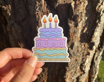 Può includere: Un adesivo di torta di compleanno a tre piani con glassa viola, blu e verde. La torta ha tre candele accese in cima.