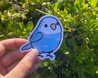 May include: A blue and white sticker of a cartoon parakeet with a pink beak and black eyes.