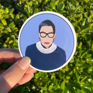 May include: A round sticker featuring a portrait of Ruth Bader Ginsburg. She is wearing a black robe, a white lace collar, and glasses. The background is a light blue color.